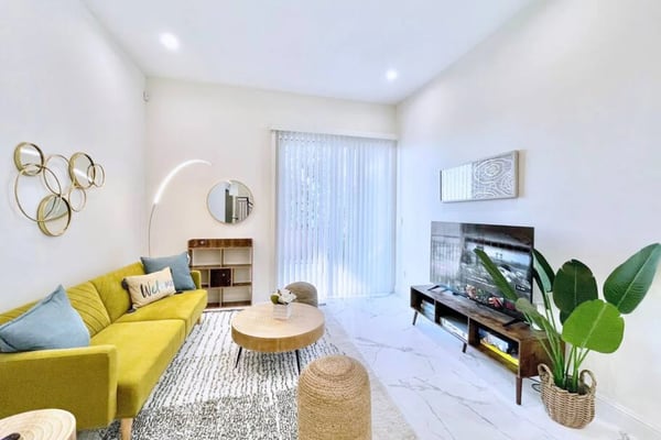 Open-plan living room with dark sofa, armchair, glass coffee tables, wall-mounted TV, and glass doors opening to backyard.