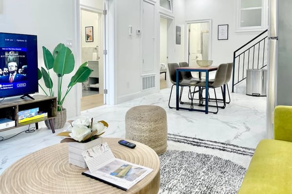 Modern open-plan living space with marble flooring, round wood table, blue dining table with gray chairs, TV, potted plant, and staircase with black railing.